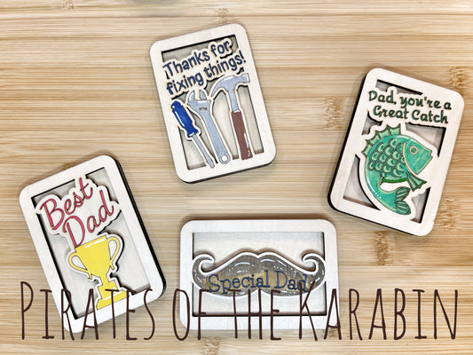 Four decorative cards with Father's Day messages on a wooden surface, featuring tools, a fish, a trophy, and a fishing reel.